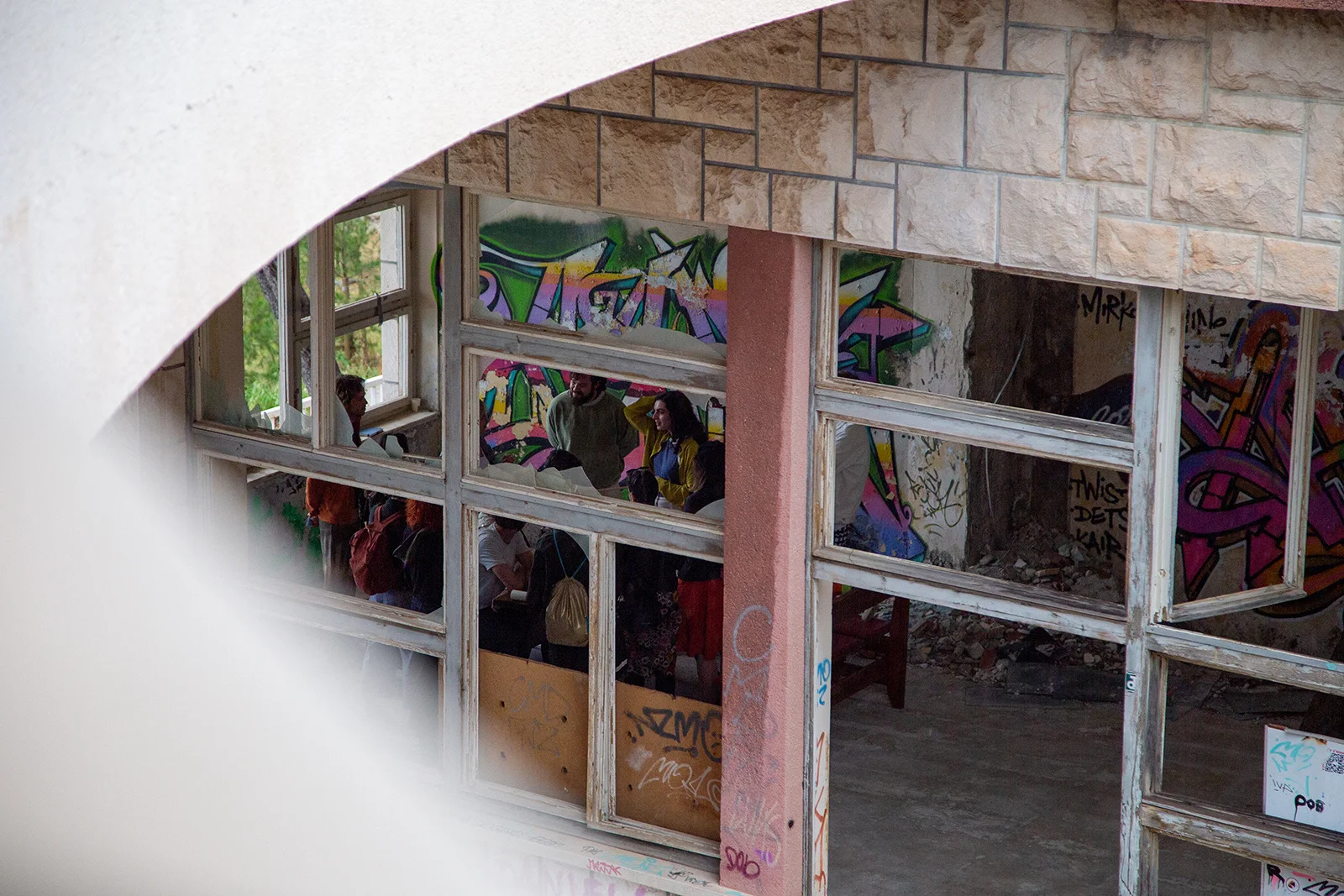 A group gathered inside a derelict modernist building, seen from outside through rows of broken window frames and graffiti-covered walls.