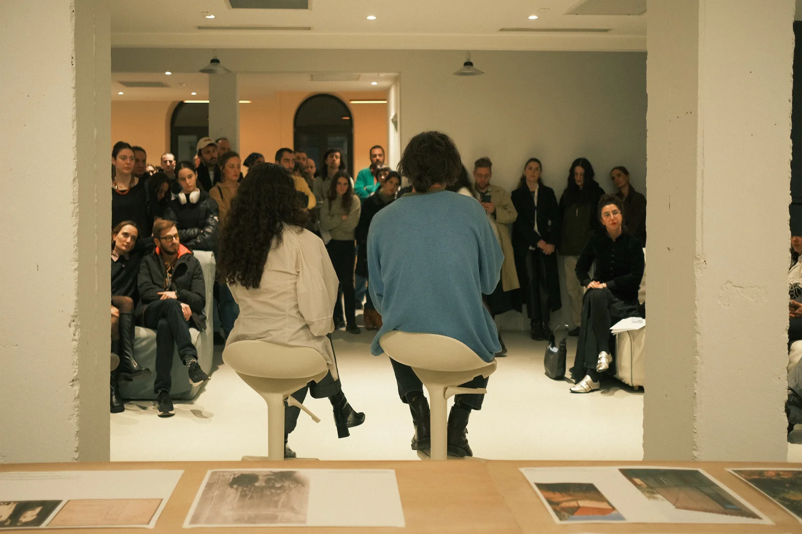 Two people seated on stools with their backs to the camera, speaking to a full room of standing and seated listeners, seen past a table of loose photographs in the foreground.