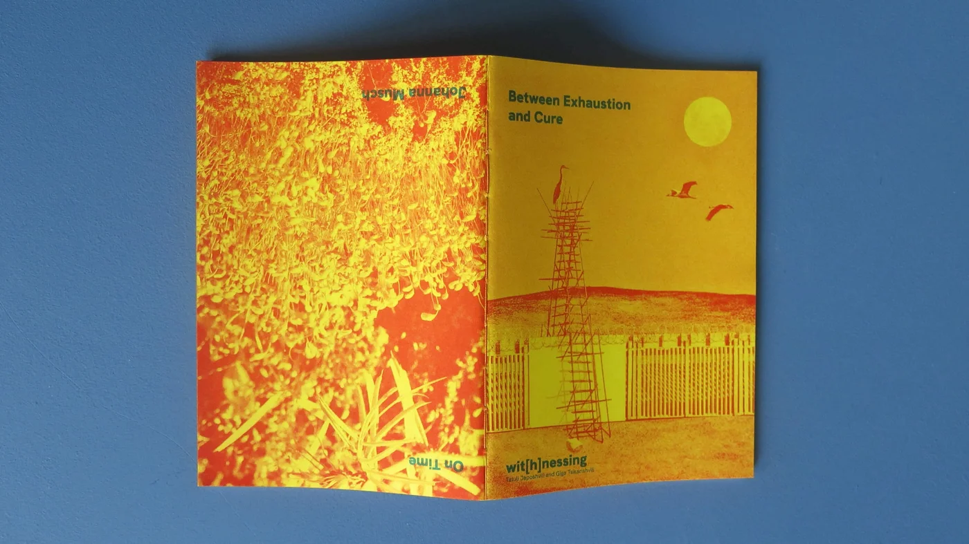 A folded riso-printed booklet in yellow and orange lying on a blue surface, its cover showing a heron on a scaffold above a fence.