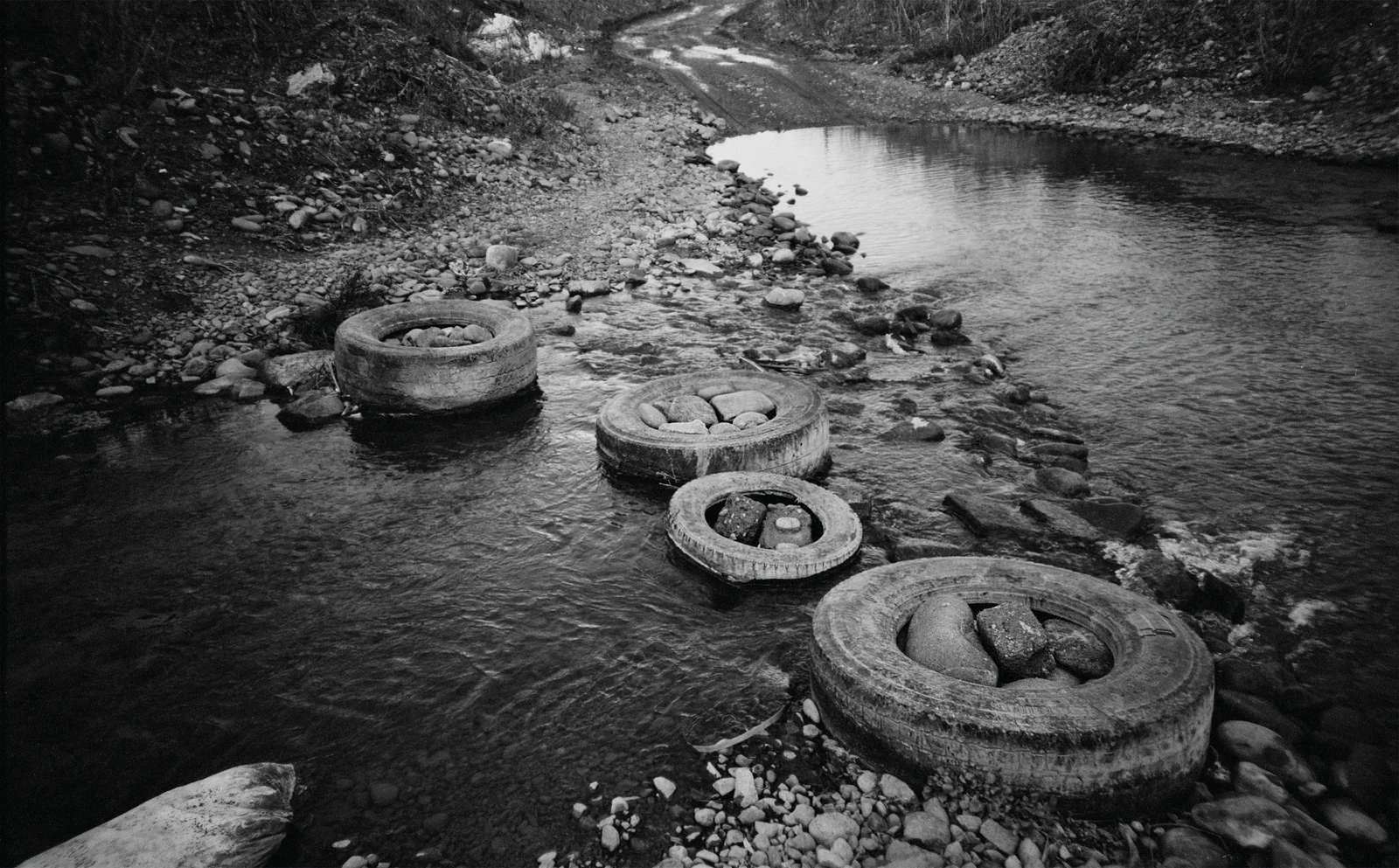 Black-and-white photograph of a dry river bed with four car tyres packed full of stones.