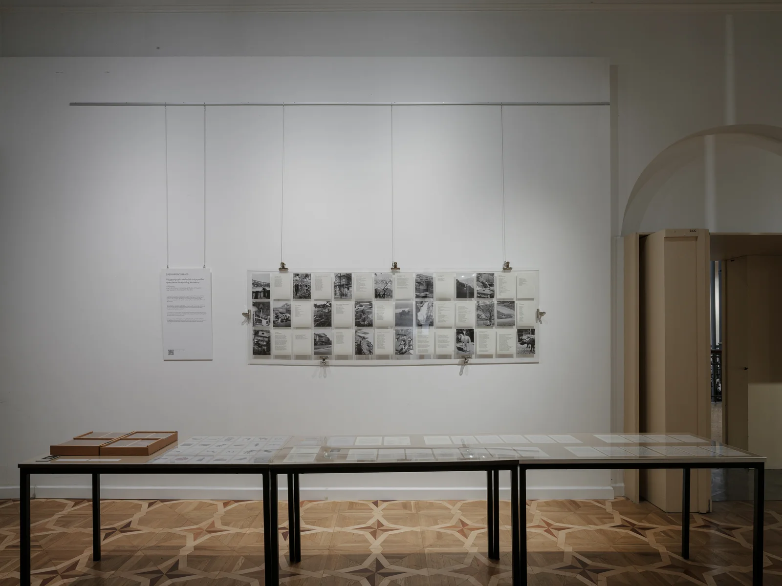 A gallery wall hung with a long grid of black-and-white photographs and captions on a suspended transparent panel, with a wall text beside it and glass-topped vitrine tables below on a patterned parquet floor.
