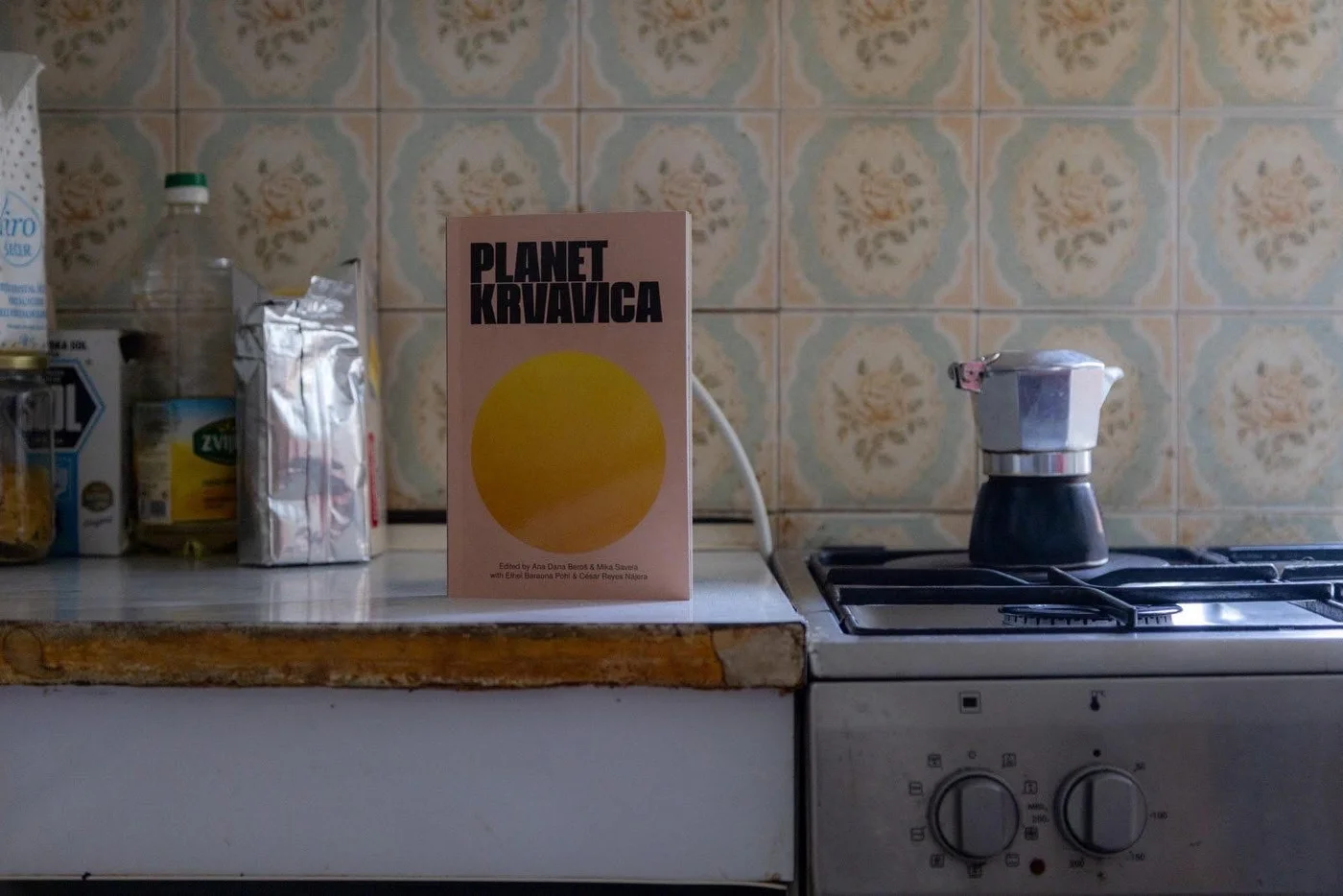 The book "Planet Krvavica" standing on a kitchen counter beside a stovetop coffee pot, against patterned floral tiles.