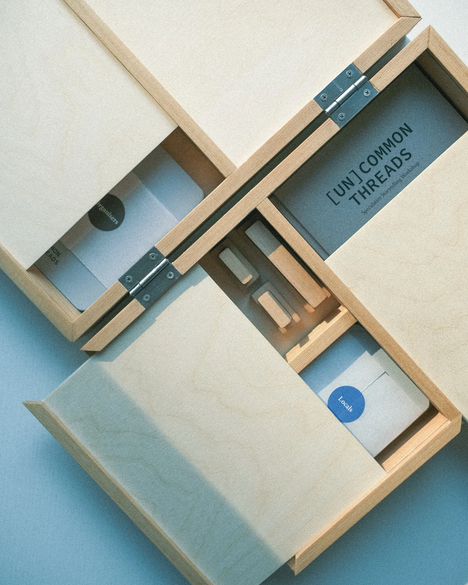 Two hinged plywood boxes lying open on a pale blue surface, holding decks of printed cards marked "Organisers" and "Locals", with a title card reading [UN]COMMON THREADS.